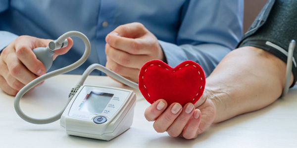 Nahaufnahme einer Blutdruckmessung: Manschette am Arm einer Person, digitales Messgerät auf dem Tisch zeigt Werte. Die Person hält ein rotes, herzförmiges Objekt in der Hand.