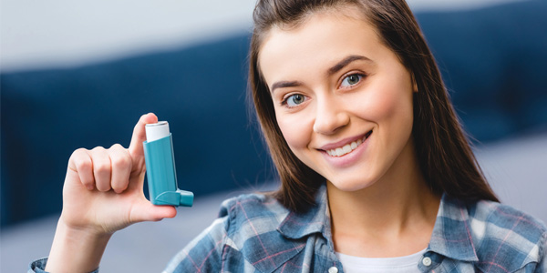 Person hält ein Inhalationsgerät in der Hand, Mundstück zeigt nach oben. Gerät wird zur Anwendung an den Mund geführt. Kleidung: kariertes Hemd. Hintergrund unscharf mit blauen Farbtönen.