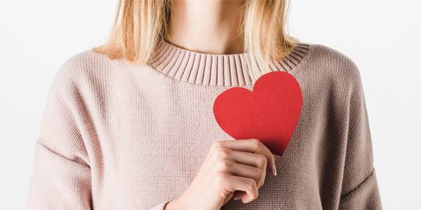 Person in hellrosa Pullover hält ein rotes Papierherz vor die Brust. Hintergrund ist weiß. Motiv erinnert visuell an Herzgesundheit.